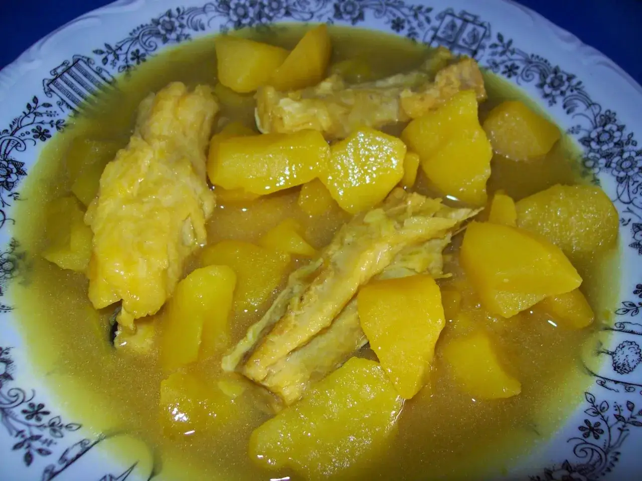 Un plato de congrio con patatas, cocinado en una salsa amarilla y servido en una vajilla con borde decorado.