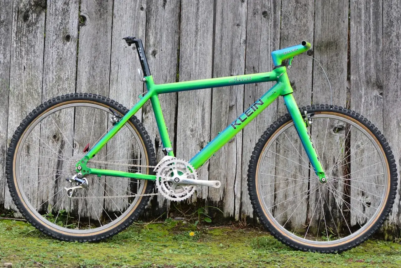 A vibrant green and blue gradient Klein Adroit mountain bike leans against a weathered wooden fence, ready for an adventure.