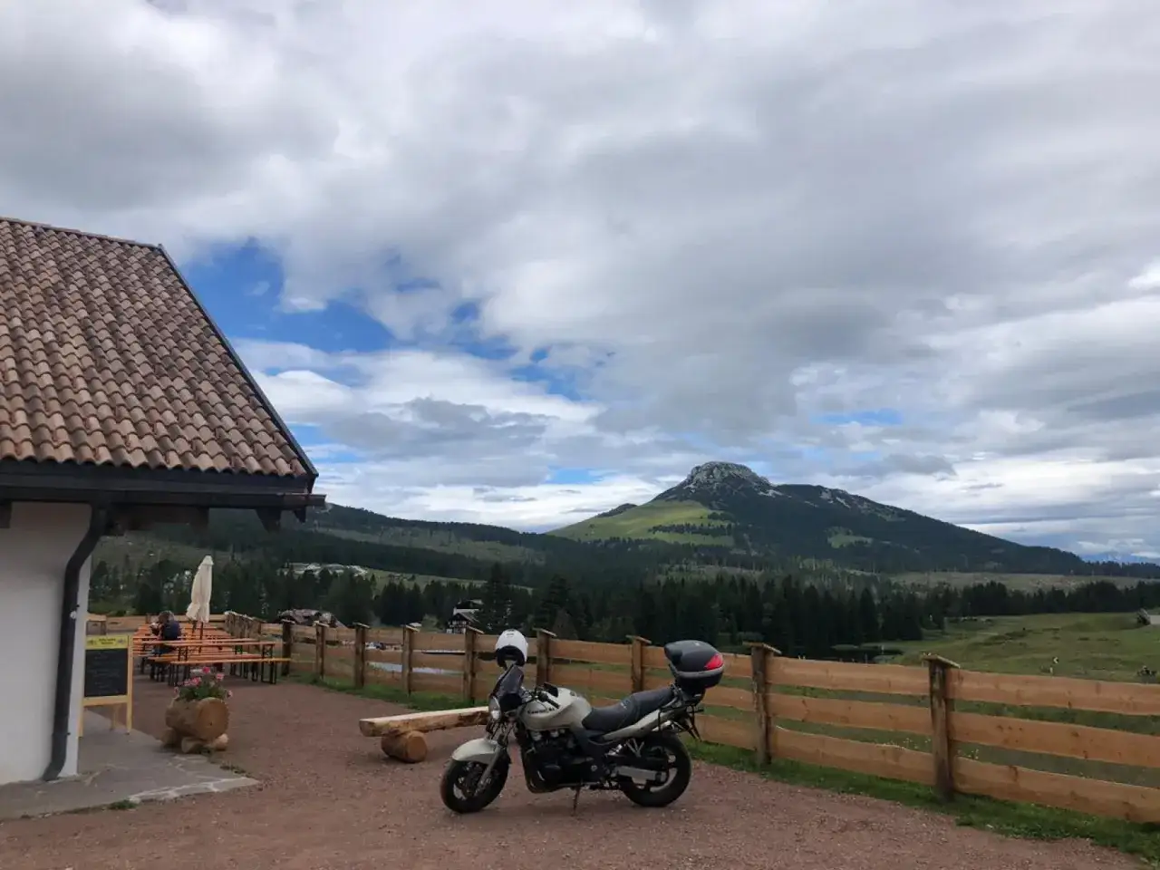 Moto parcheggiata vicino a un rifugio con tetto in tegole rosse, sullo sfondo le montagne e le malghe di Passo Lavazè.