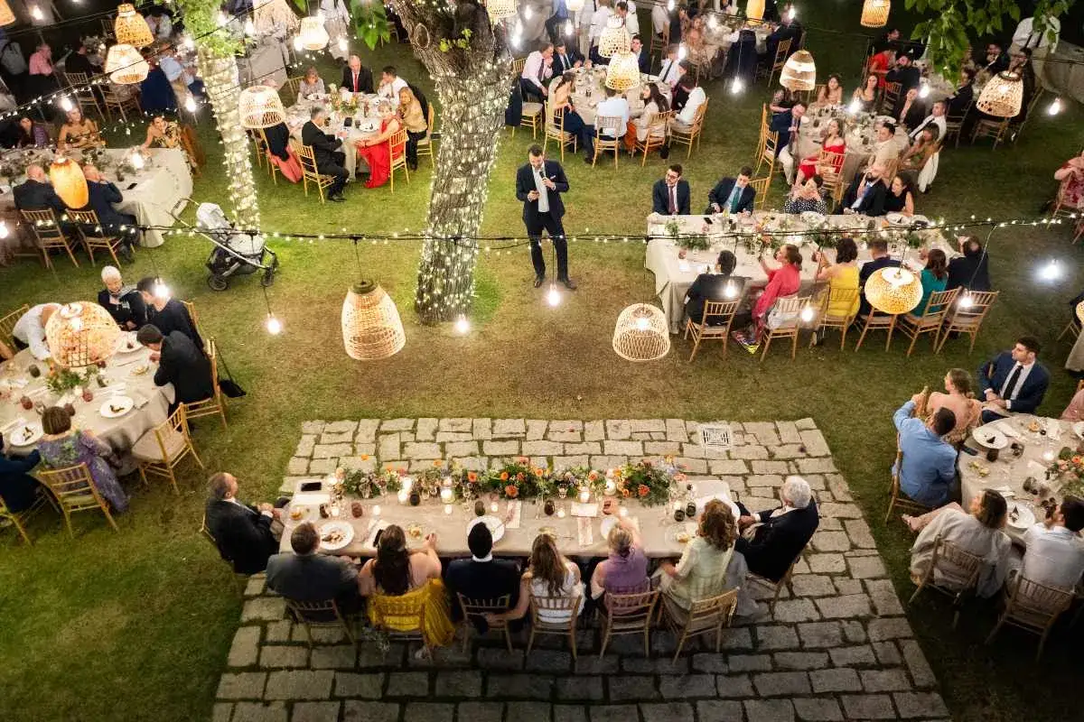 Una elegante mesa presidencial de boda, iluminada por luces c&aacute;lidas, con invitados disfrutando de la velada al aire libre.