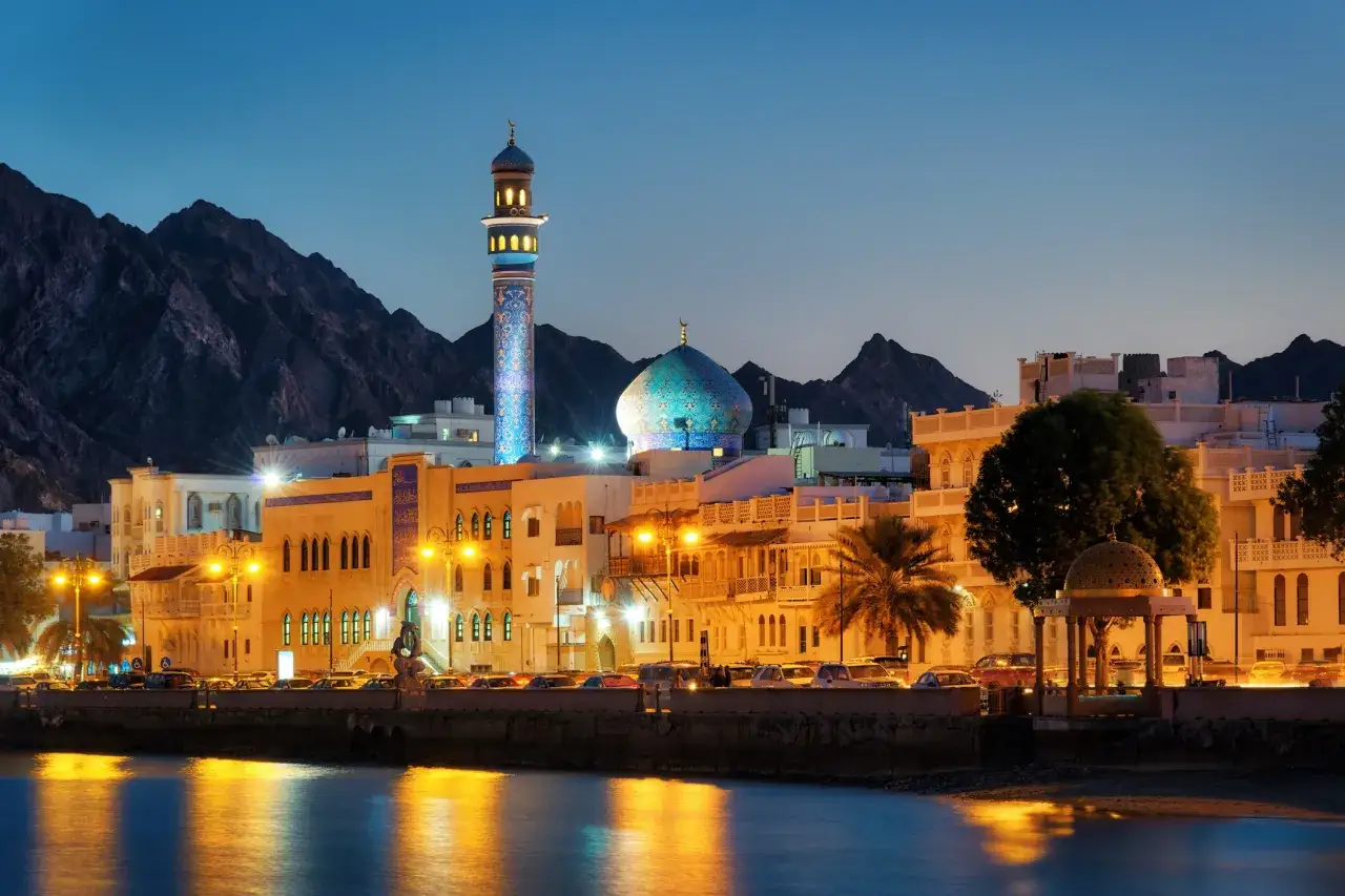 Abendliche Skyline von Maskat, Oman, mit einer Moschee, deren Minarett und Kuppel beleuchtet sind, vor majest&auml;tischen Bergen.