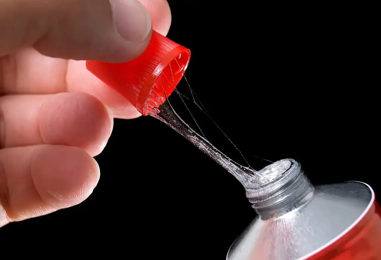 Main d'une personne tenant le bouchon rouge d'un tube de colle super glue, avec des fils de colle s'&eacute;tirant. Attention, colle super glue sur les doigts dangereux.