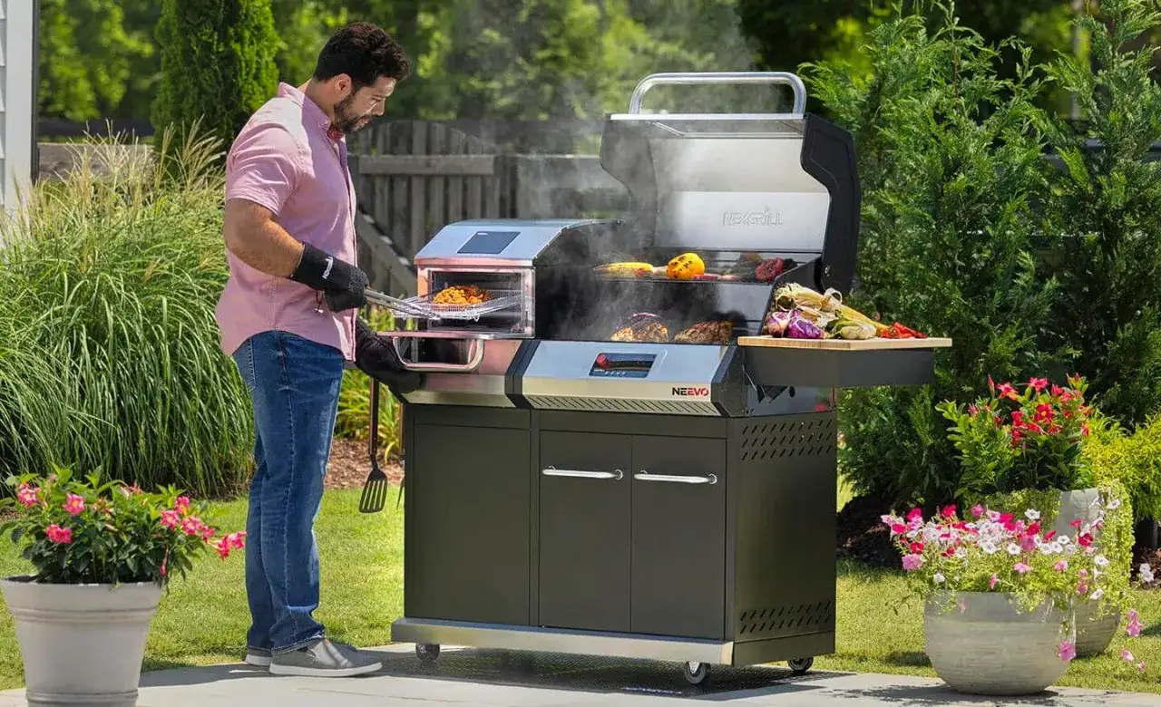Mężczyzna w różowej koszuli przygotowuje jedzenie na nowoczesnym grillu. Widać dymiące mięso, kukurydzę i warzywa. To jest właśnie grill!