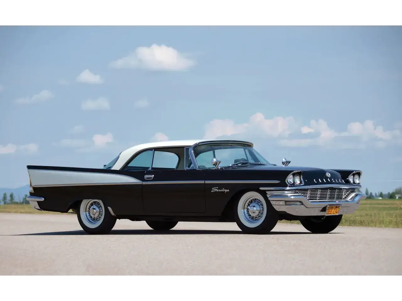 Svart Chrysler Saratoga med vit tak och vita däcksidor, parkerad på en asfalterad väg under en molnig himmel.