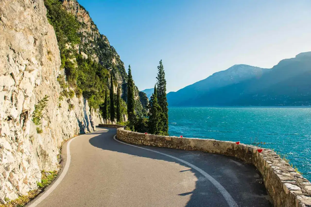 Scenic Italian lakeside road, perfect for exploring after checking renting a car in Italy requirements. Mountains and cypress trees line the turquoise water.