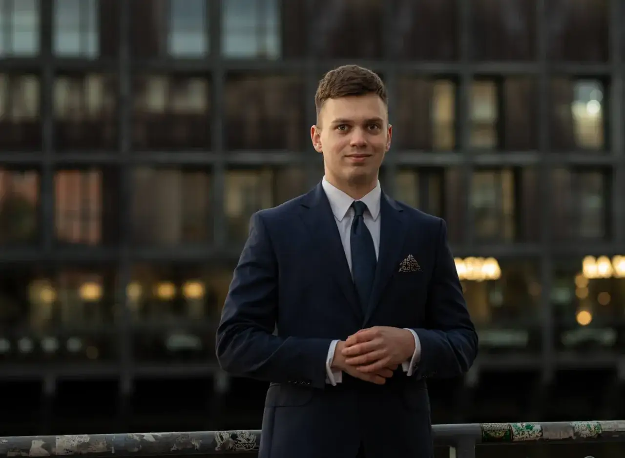 Młody mężczyzna w granatowym garniturze i krawacie stoi przed budynkiem.