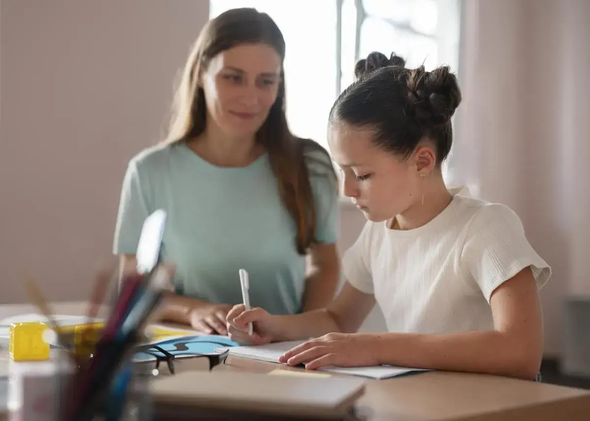 Ni&ntilde;a concentrada escribiendo, con una mujer observ&aacute;ndola. Una escena de apoyo en la oposici&oacute;n orientaci&oacute;n educativa.
