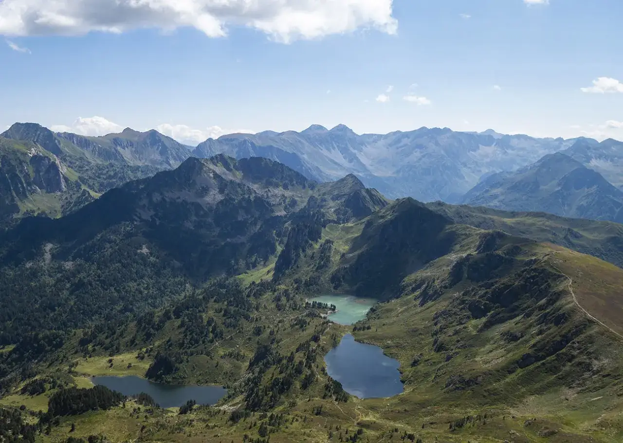 Vue aérienne des Pyrénées avec l'étang de Rabassoles, un lac bleu profond niché entre des montagnes verdoyantes et des pics rocheux sous un ciel bleu clair.