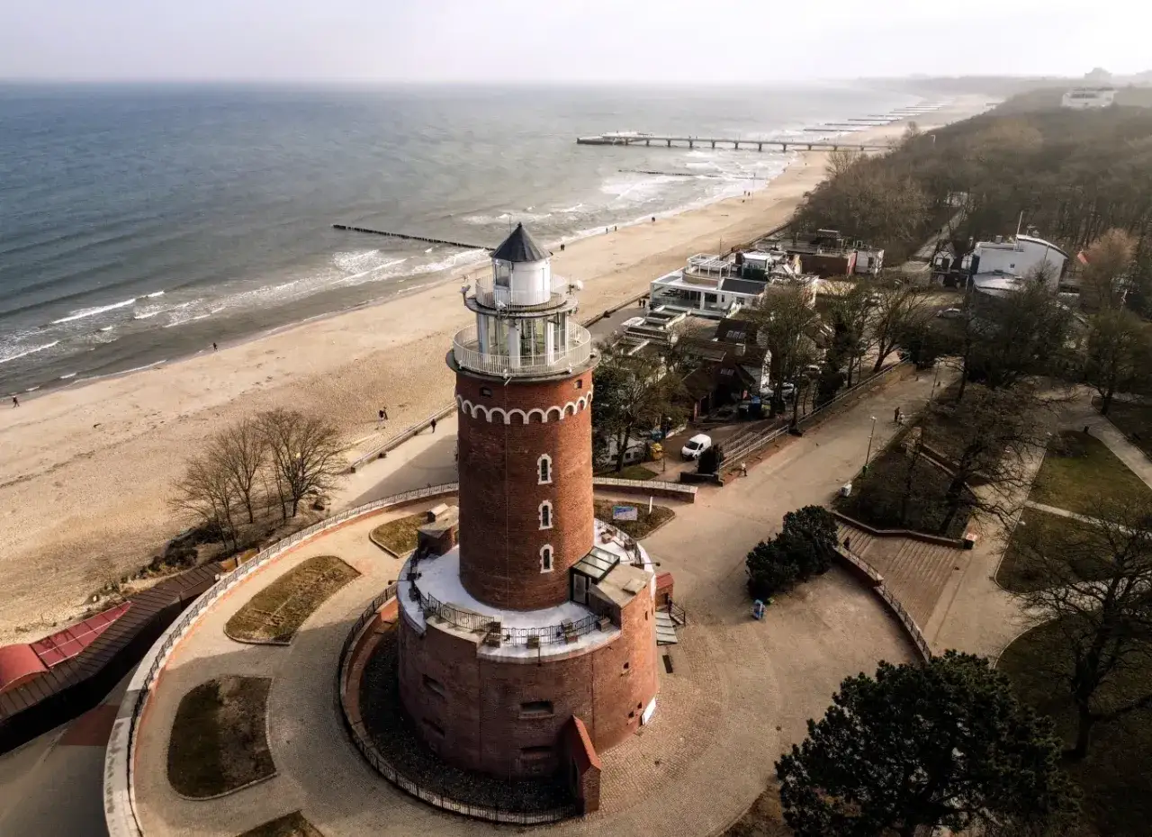 Ceglana latarnia morska góruje nad piaszczystą plażą i morzem. W oddali widać molo i zabudowania.