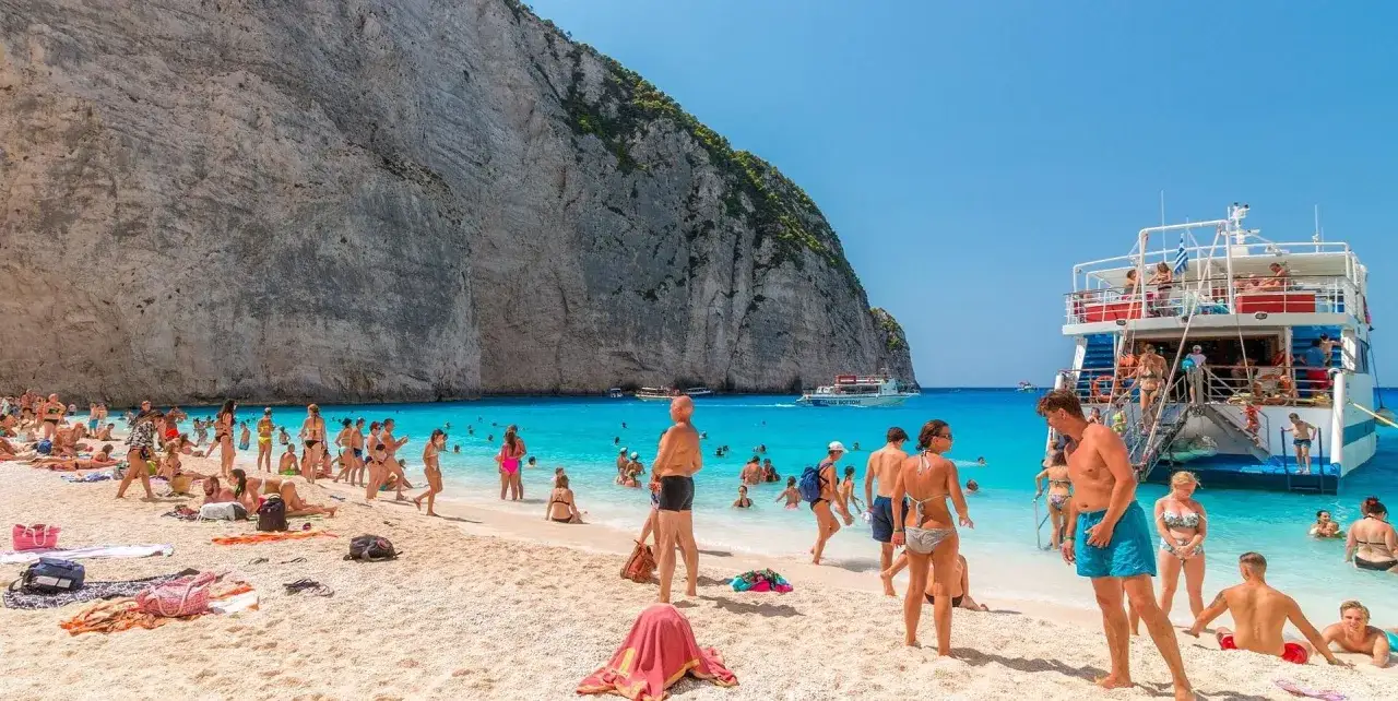 Tłum ludzi na plaży Navagio, znanej jako zatoka wraku, cieszy się słońcem i turkusową wodą. Statek wycieczkowy cumuje blisko brzegu.