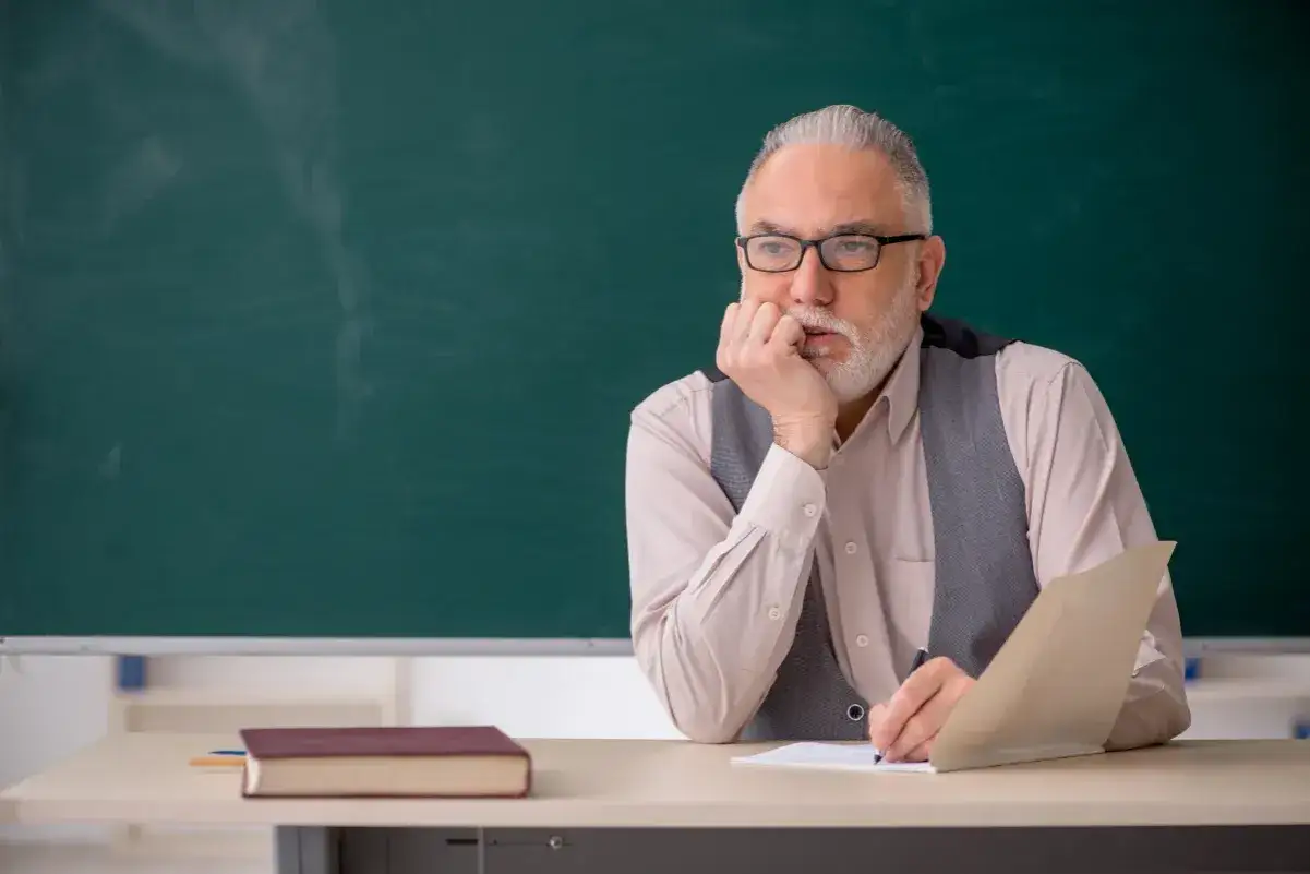 Starszy nauczyciel zamyślony nad zeszytem, przygotowanie pedagogiczne. Obok książka i zielona tablica.