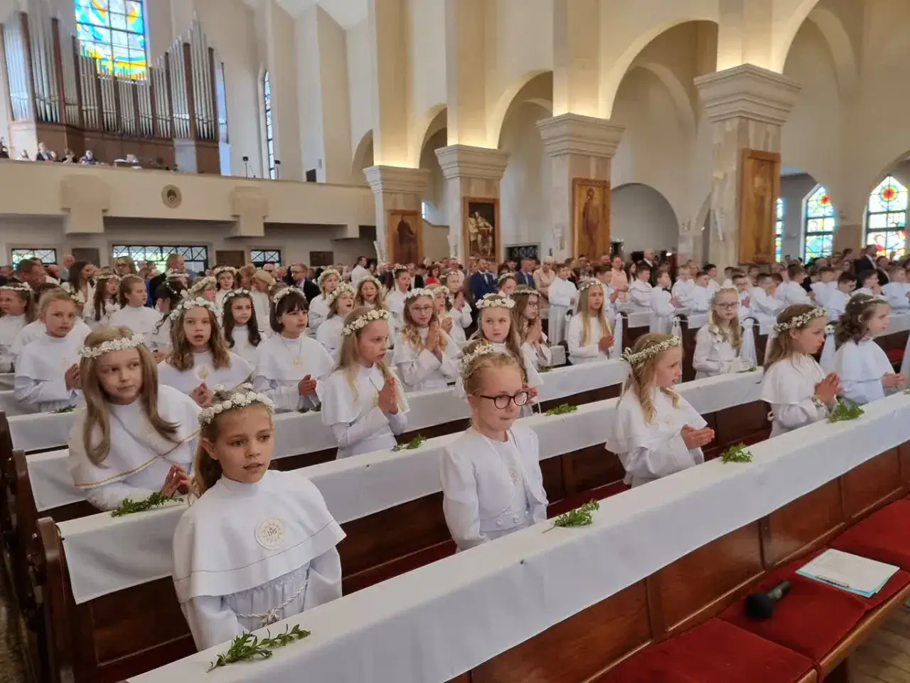 Dzieci w białych strojach, z wiankami na głowach, siedzą w kościele, czekając na moment, kiedy sa komunie.