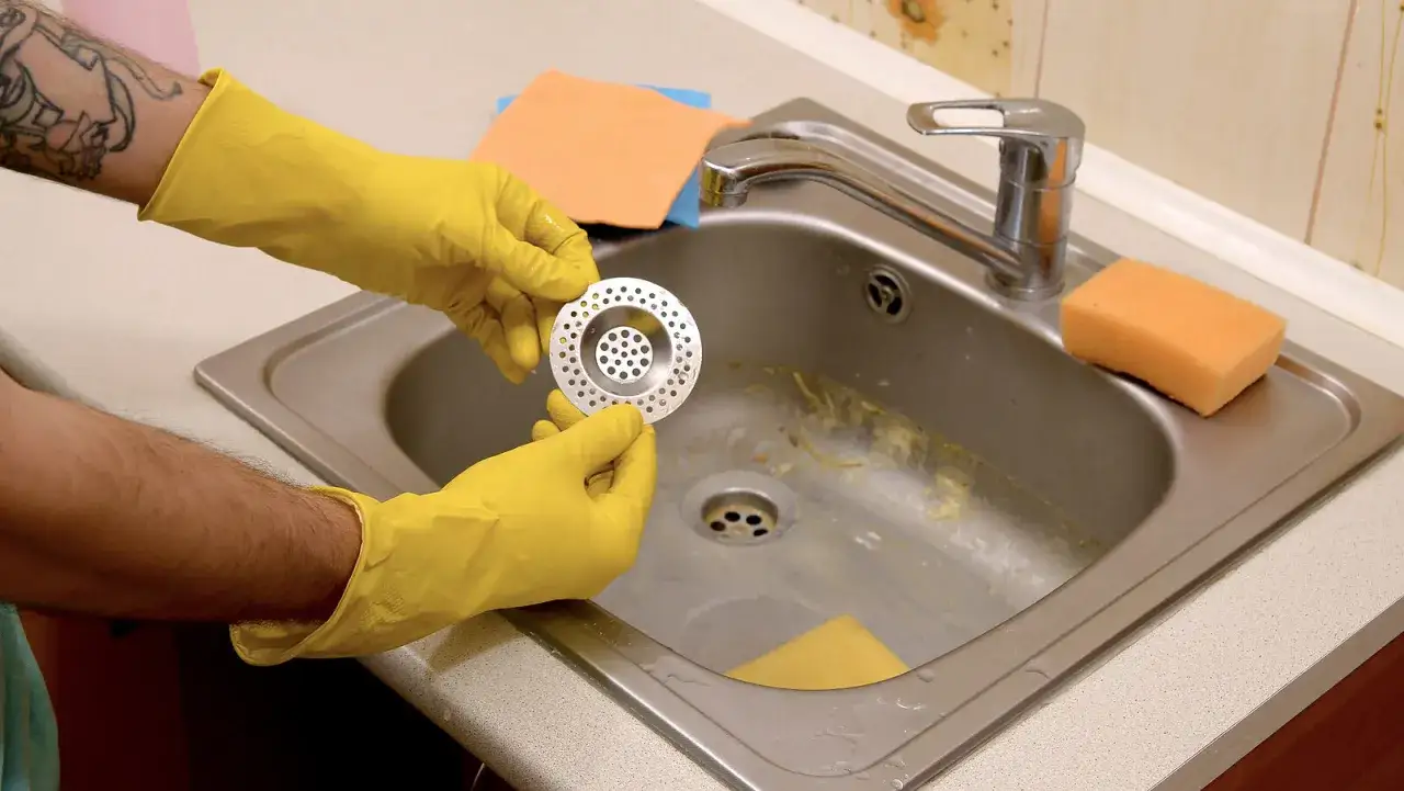 Manos con guantes amarillos sostienen un colador para el esquema del desague de la cocina. Hay una esponja naranja y paños cerca.