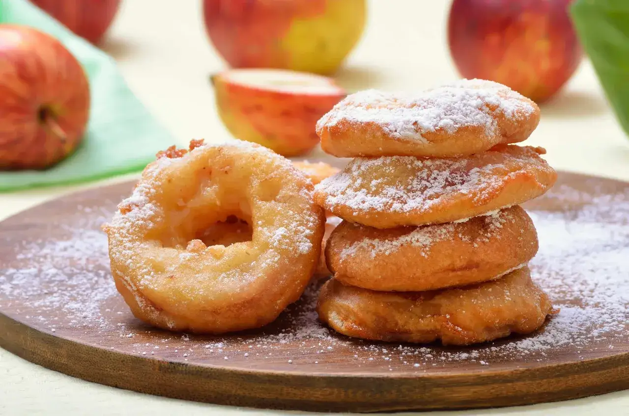 Délicieux beignets aux pommes à lancienne, saupoudrés de sucre glace. Des pommes fraîches en arrière-plan rappellent la douceur de cette recette.