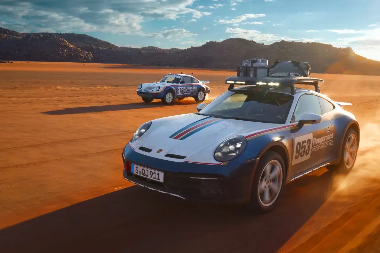 Två Porsche 911, en modern och en klassisk, kör i öknen. Den moderna Porsche safari-bilen har takbox och extraljus.
