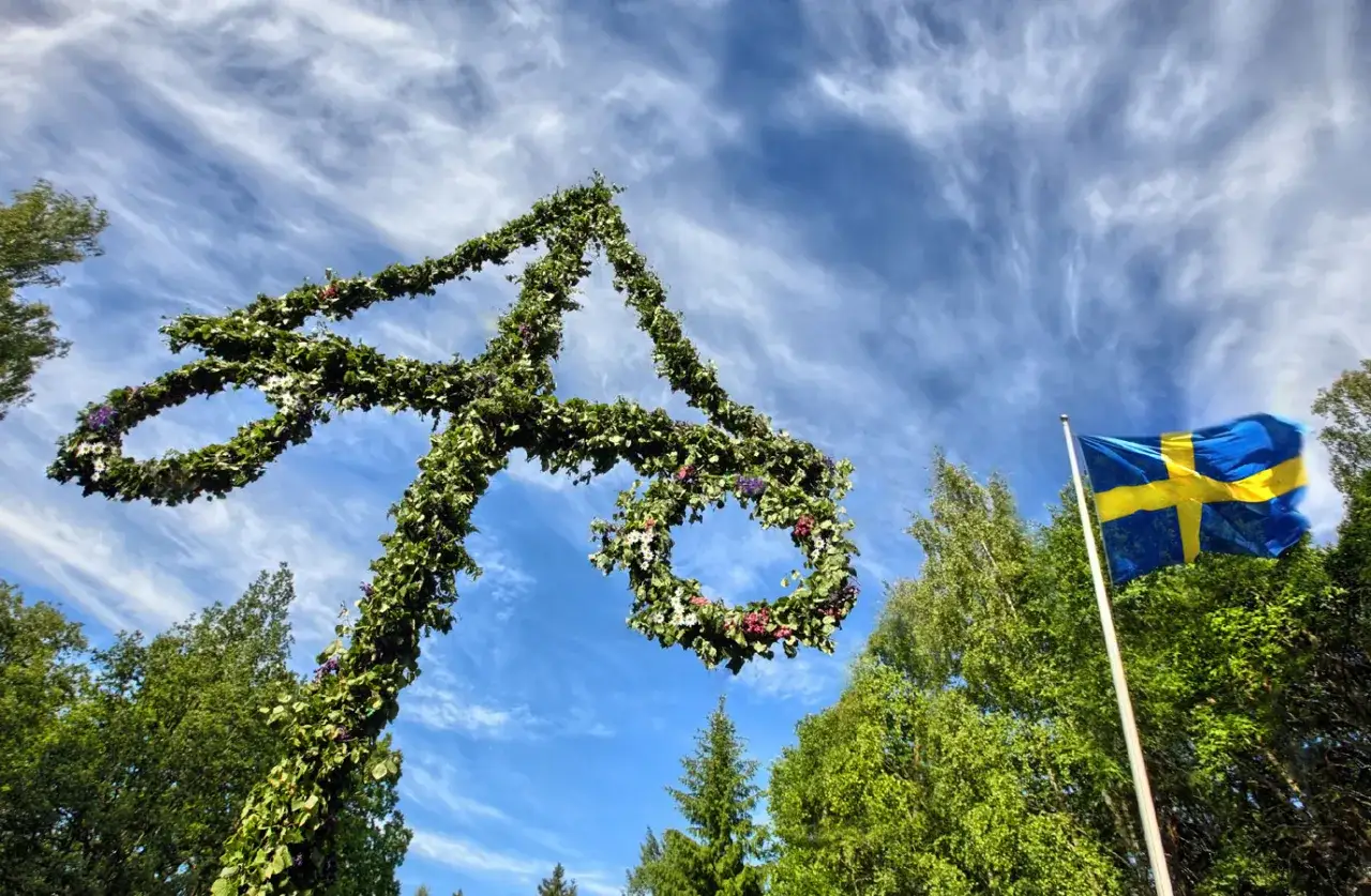 Blomsterprydd midsommarstång och svensk flagga mot en blå himmel med moln. En vacker scen som visar vilka firar midsommar.