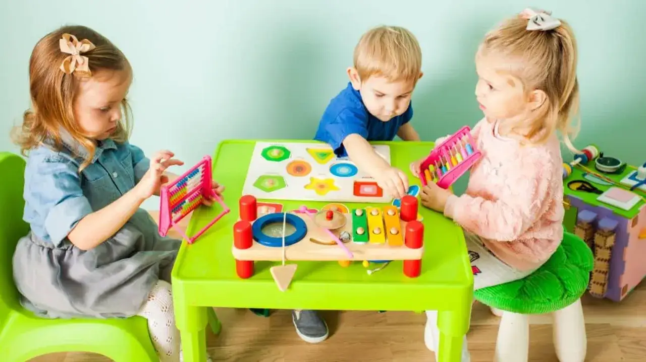 Dzieci bawią się zabawkami edukacyjnymi przy zielonym stoliku. Czy żłobek jest płatny? To pytanie nurtuje wielu rodziców.
