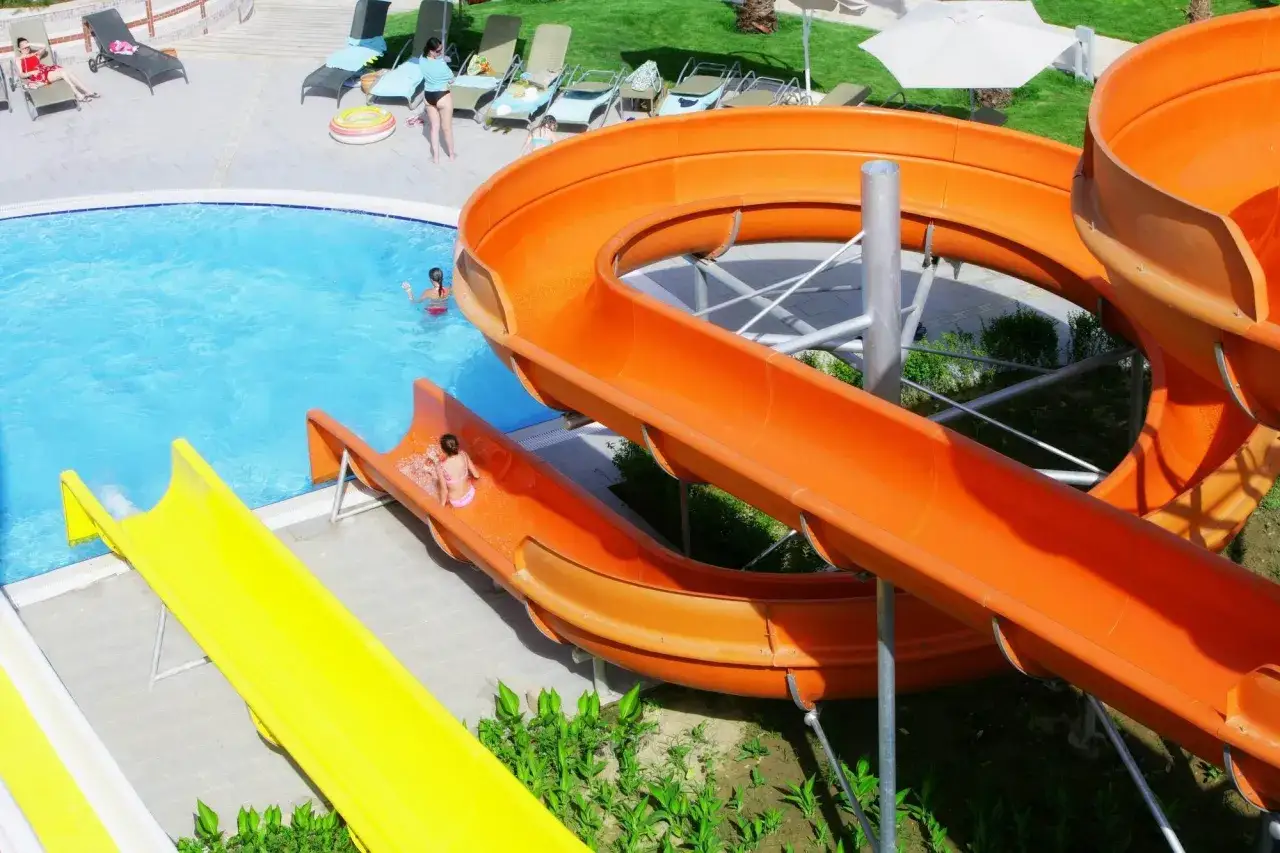 Dzieci zjeżdżają ze zjeżdżalni wodnych w basenie. Wiele osób odpoczywa na leżakach. **Mukarnas spa resort opinie** są pozytywne.