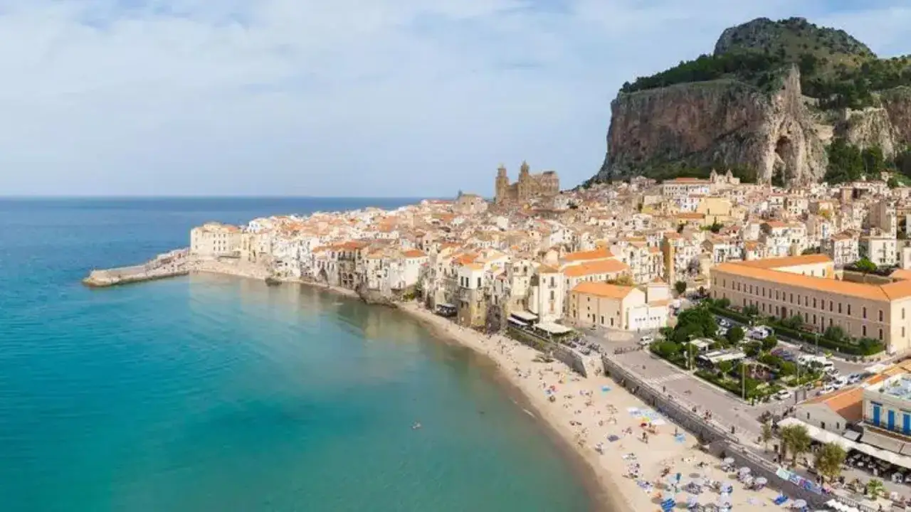 Vue aérienne d'une ville pittoresque en **Italie, bord de mer**, avec une plage animée et une mer turquoise. Un rocher imposant domine le paysage.