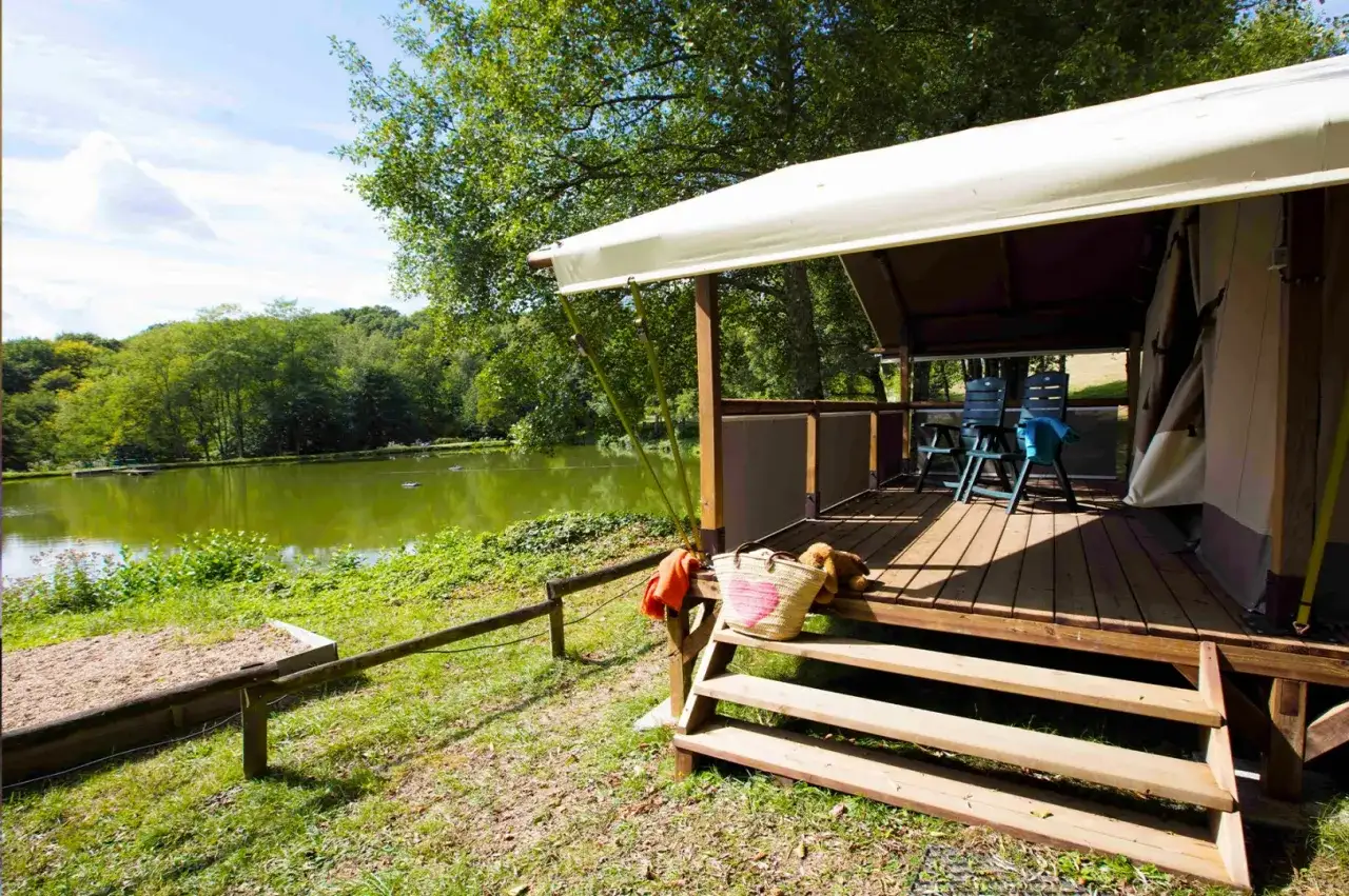 Un camping &eacute;co-lodge avec une terrasse en bois donnant sur un lac paisible. Des chaises et un sac de plage sont pr&ecirc;ts pour la d&eacute;tente.