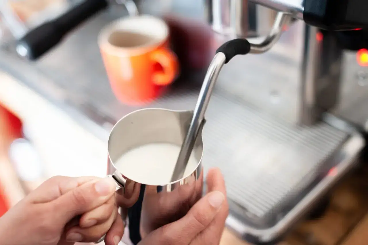 Mani che preparano un cappuccino senza schiuma, con una tazza arancione sullo sfondo.