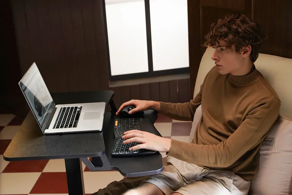 Junger Mann mit Laptop und Tastatur am Bett. Mit Tennisarm darf man mit diesem ergonomischen Tisch arbeiten.