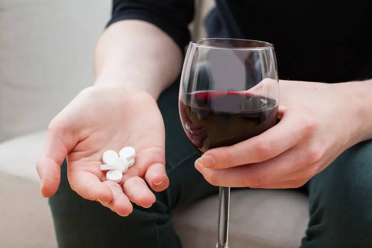 Dłoń trzyma białe tabletki, obok kieliszek czerwonego wina. Seronil a alkohol to niebezpieczne połączenie.