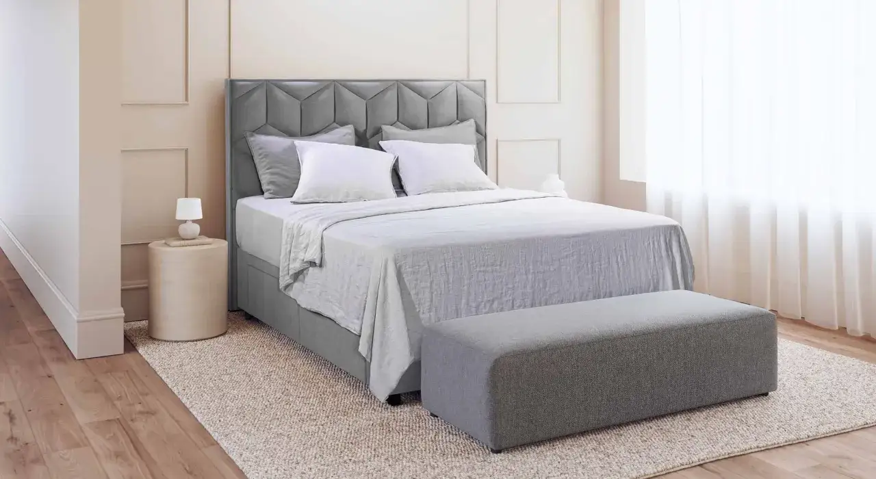 Lit de dimension queen size, avec tête de lit capitonnée grise, draps blancs et banc assorti.