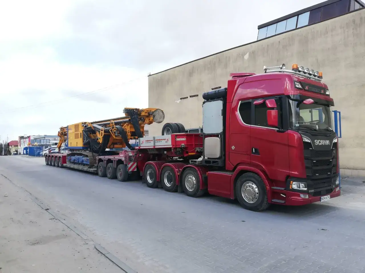 Czerwona Scania z naczepą transportuje żółtą wiertnicę. To prawdziwa przesyłka dużych gabarytów, gotowa do drogi.