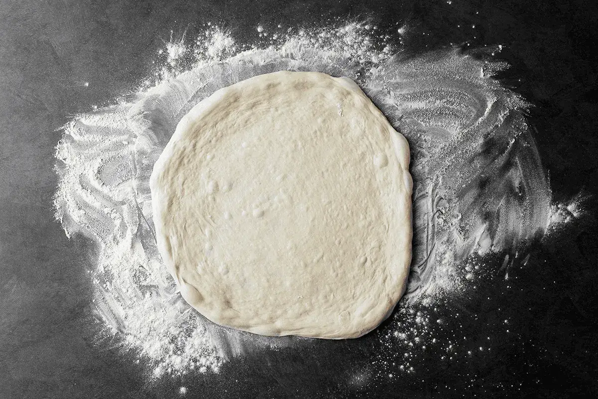 Okrągłe ciasto na pizzę na cienkim cieście, otoczone mąką na ciemnym tle. Gotowe do pieczenia.