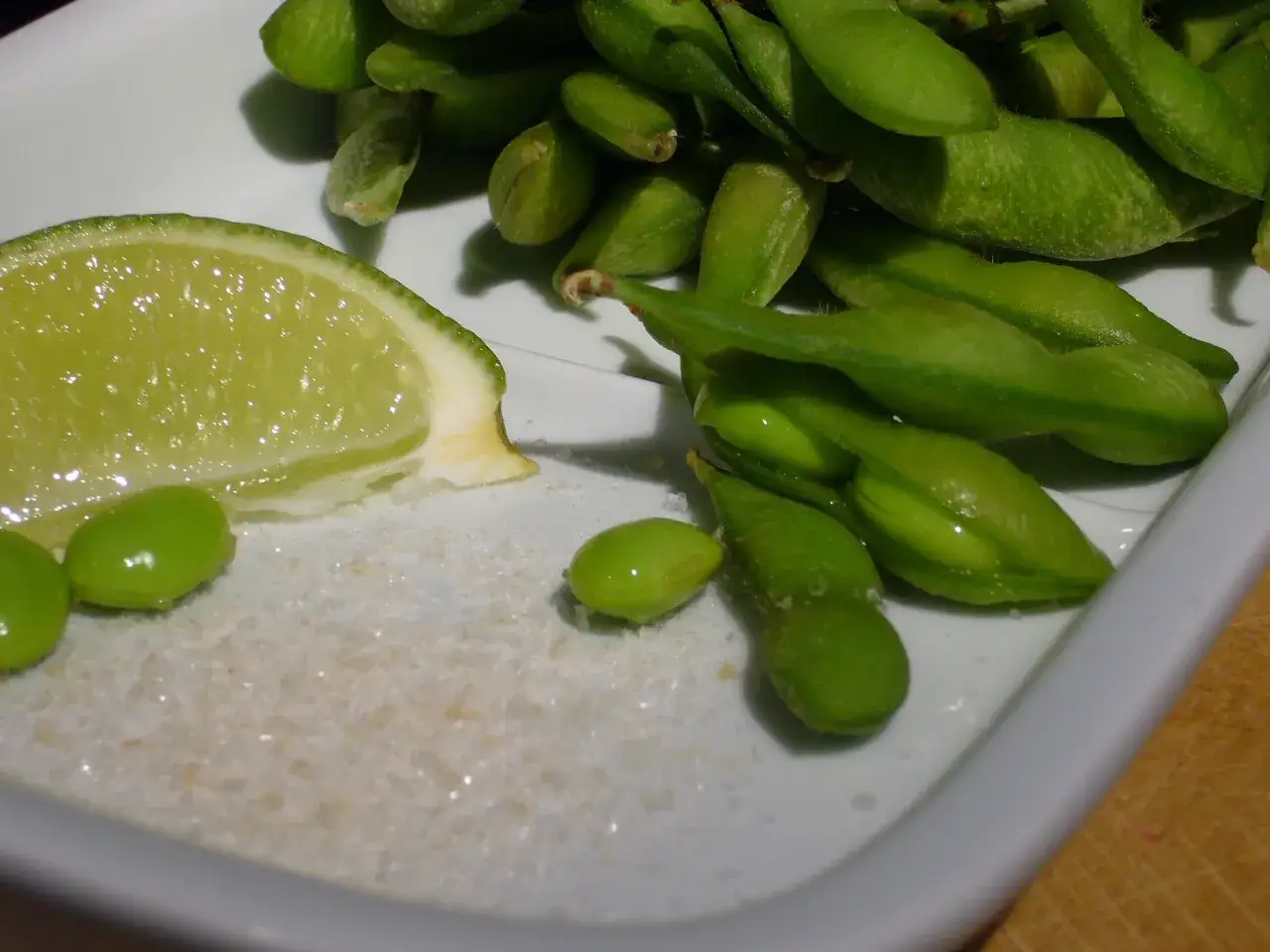 Gr&uuml;ne Edamame-Schoten, eine Limettenspalte und Salz auf einem Teller. Was sind Edamame? Junge Sojabohnen, die man so genie&szlig;t.