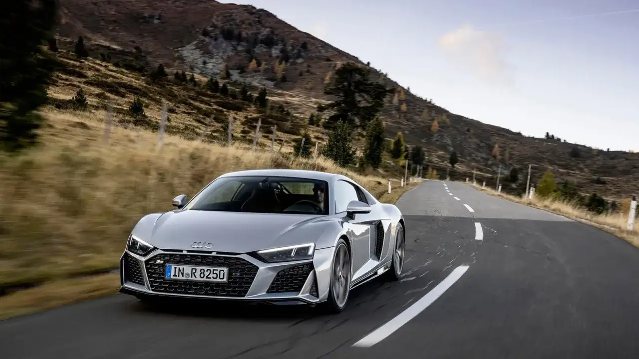 Un Audi R8 plateado recorre una carretera de montaña. El coche, de donde es Audi, se ve deportivo y elegante.