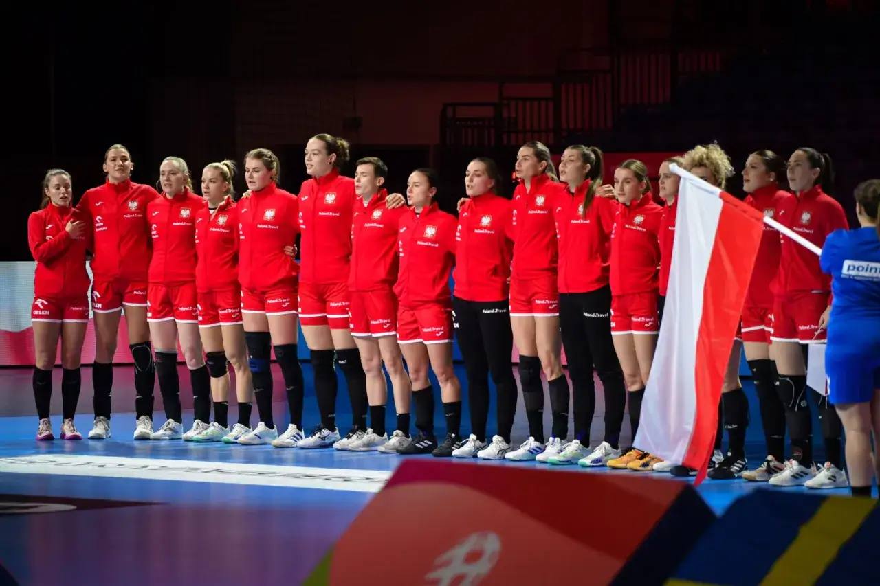 Reprezentacja Polski w piłce ręcznej kobiet stoi w rzędzie, gotowa do meczu. Biało-czerwona flaga symbolizuje dumę narodową.