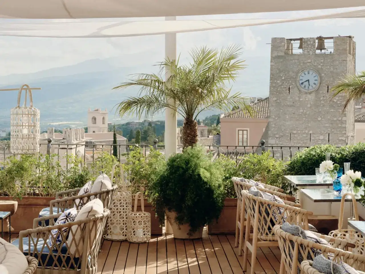 Terrazza con sedie in vimini e vista panoramica su Taormina. Il **bar Timeo Taormina** offre un'atmosfera rilassante con palme e lanterne.