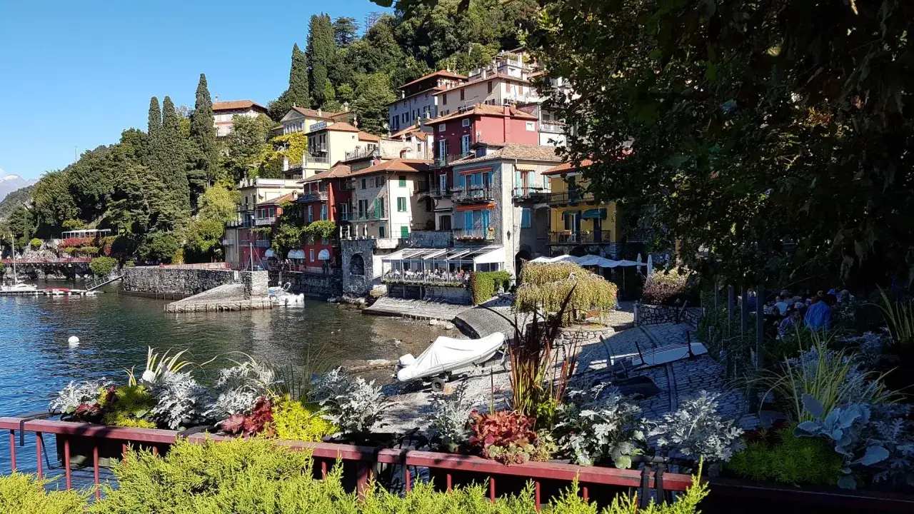 Bunte H&auml;user am Seeufer, wie como see, mit Terrassen und B&auml;umen. Ein Boot liegt am Ufer.