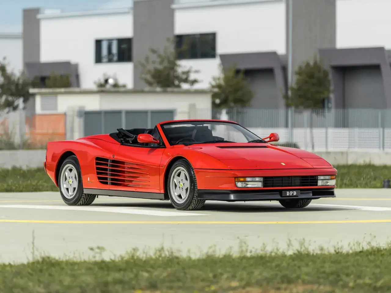 Ein roter Ferrari Testarossa Spider steht auf einem Parkplatz vor modernen Geb&auml;uden.