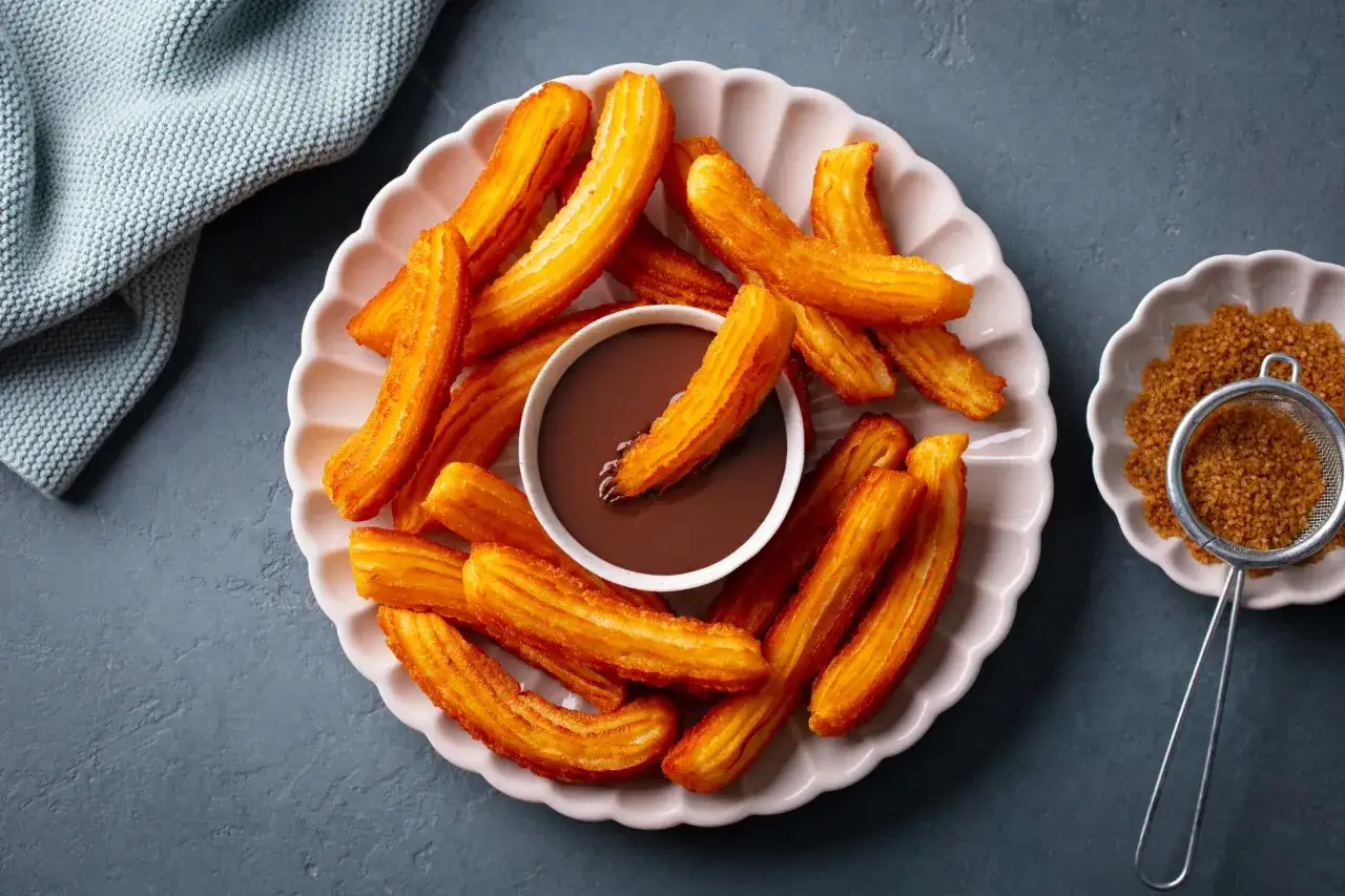 Une assiette de churros dorés et croustillants, parfaits pour une recette facile. Un bol de chocolat chaud les accompagne.
