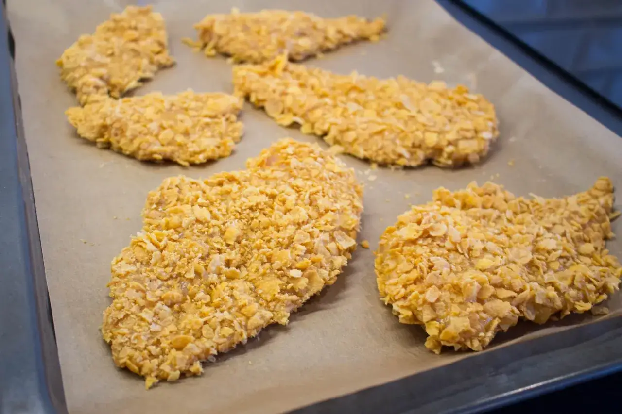 Flera bitar cornflakes kyckling ligger p&aring; ett bakpl&aring;tspapper, redo att tillagas.