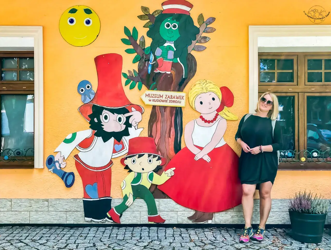 Kobieta pozuje przy kolorowej ścianie Muzeum Zabawek w Kudowie-Zdroju, Dolny Śląsk. Bajkowe postacie i słońce tworzą radosną atmosferę.