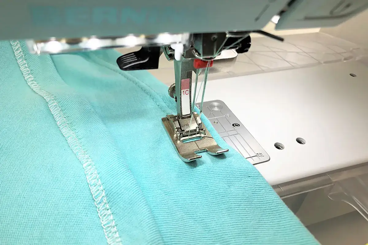 A sewing machine with a twin needle stitches a hem on turquoise fabric. This shows how you can backstitch with a twin needle for a clean finish.