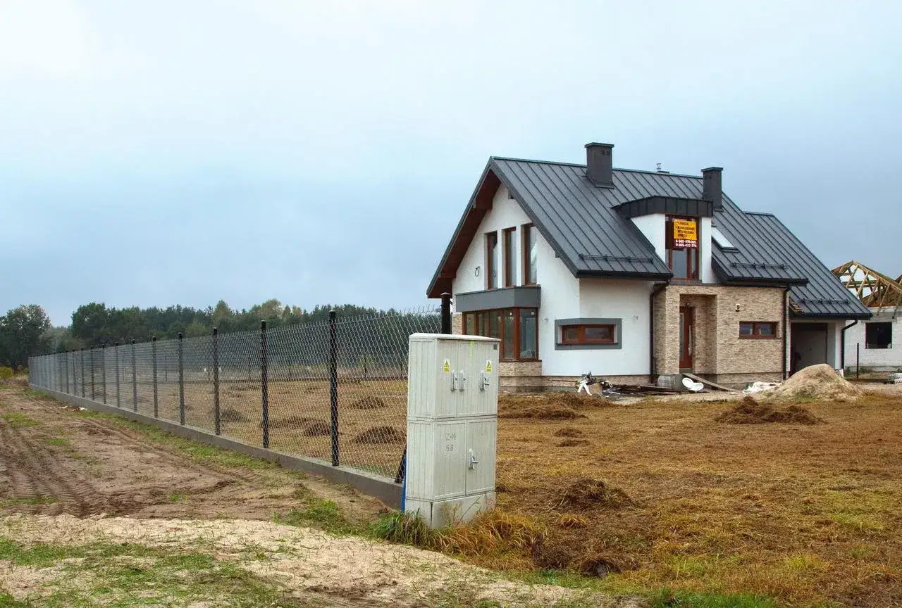 Podłączenie prądu do działki bez domu. Obok ogrodzenia stoi skrzynka elektryczna, a w tle widać budowany dom.