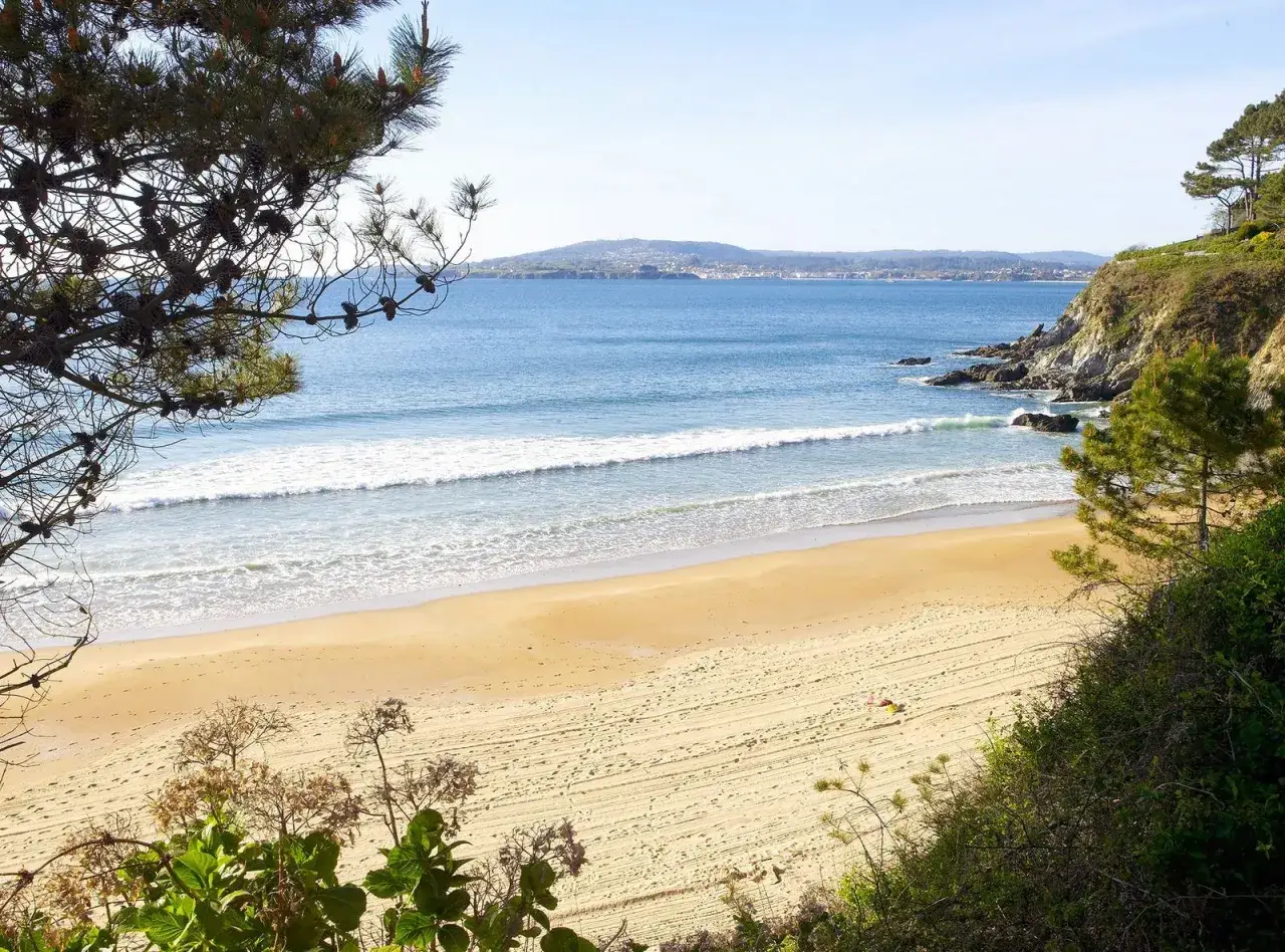 Una persona descansa en la arena dorada de la playa ber, con olas suaves rompiendo en la orilla y un paisaje costero al fondo.