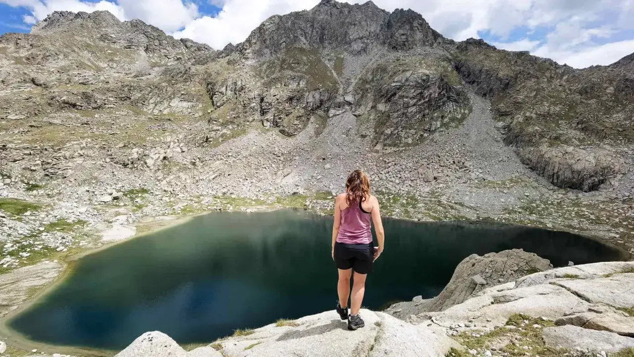 Mujer admira un estany gento rodeado de monta&ntilde;as rocosas y un cielo azul con nubes.