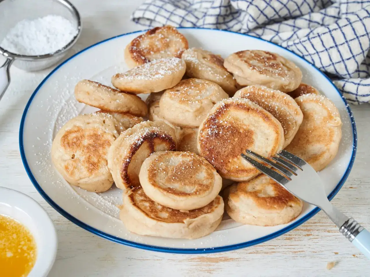 Ein Teller voller goldbrauner Poffertjes, bestäubt mit Puderzucker. Ein Gabel liegt bereit, um dieses köstliche Poffertjes Rezept zu genießen.