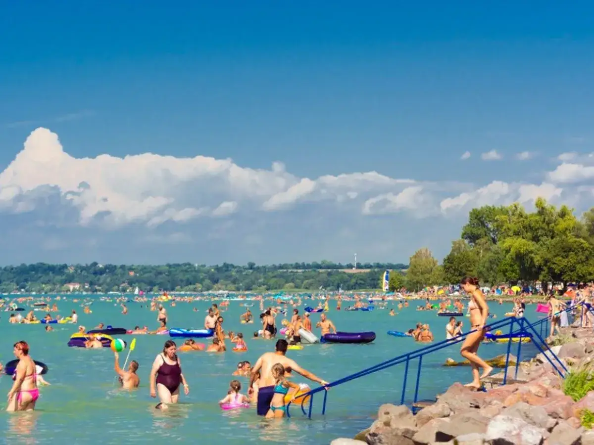 Tłum ludzi cieszy się słońcem i wodą na zatłoczonej **balaton plaży**. Dzieci pływają na dmuchanych zabawkach, dorośli relaksują się w wodzie.