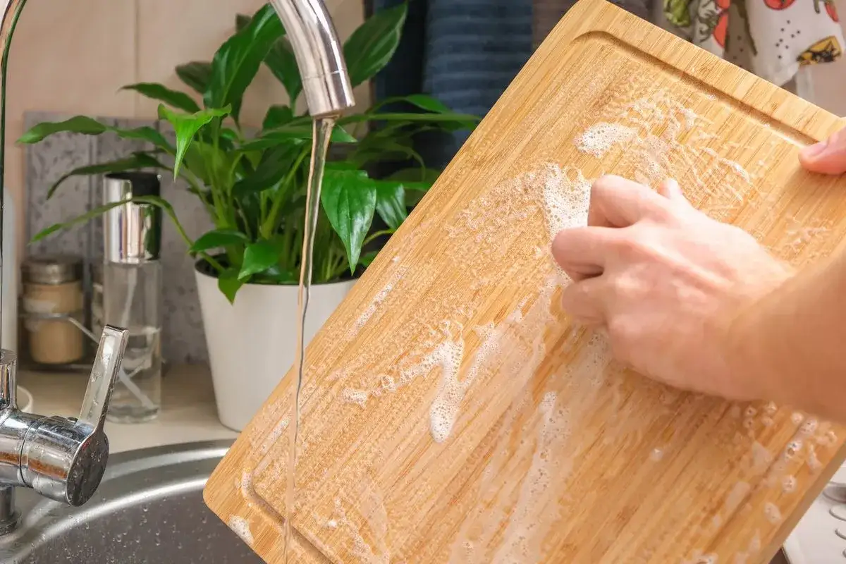 Nettoyer planche &agrave; d&eacute;couper en bois avec du bicarbonate de soude et de l'eau. Une main frotte la surface savonneuse sous le robinet.