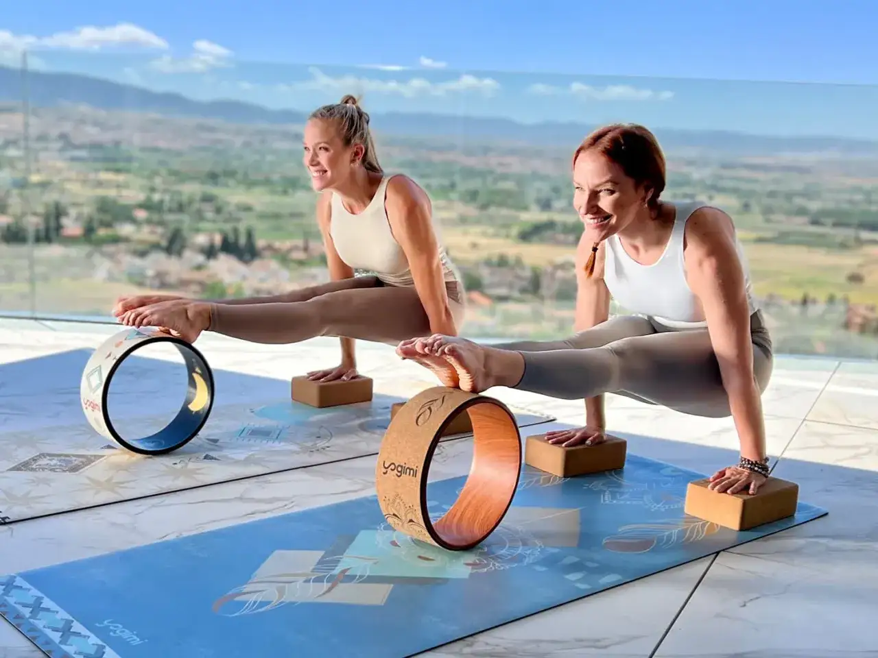 Dos mujeres practican posturas de yoga con una rueda de yoga, apoyadas en bloques, con un paisaje montañoso de fondo.