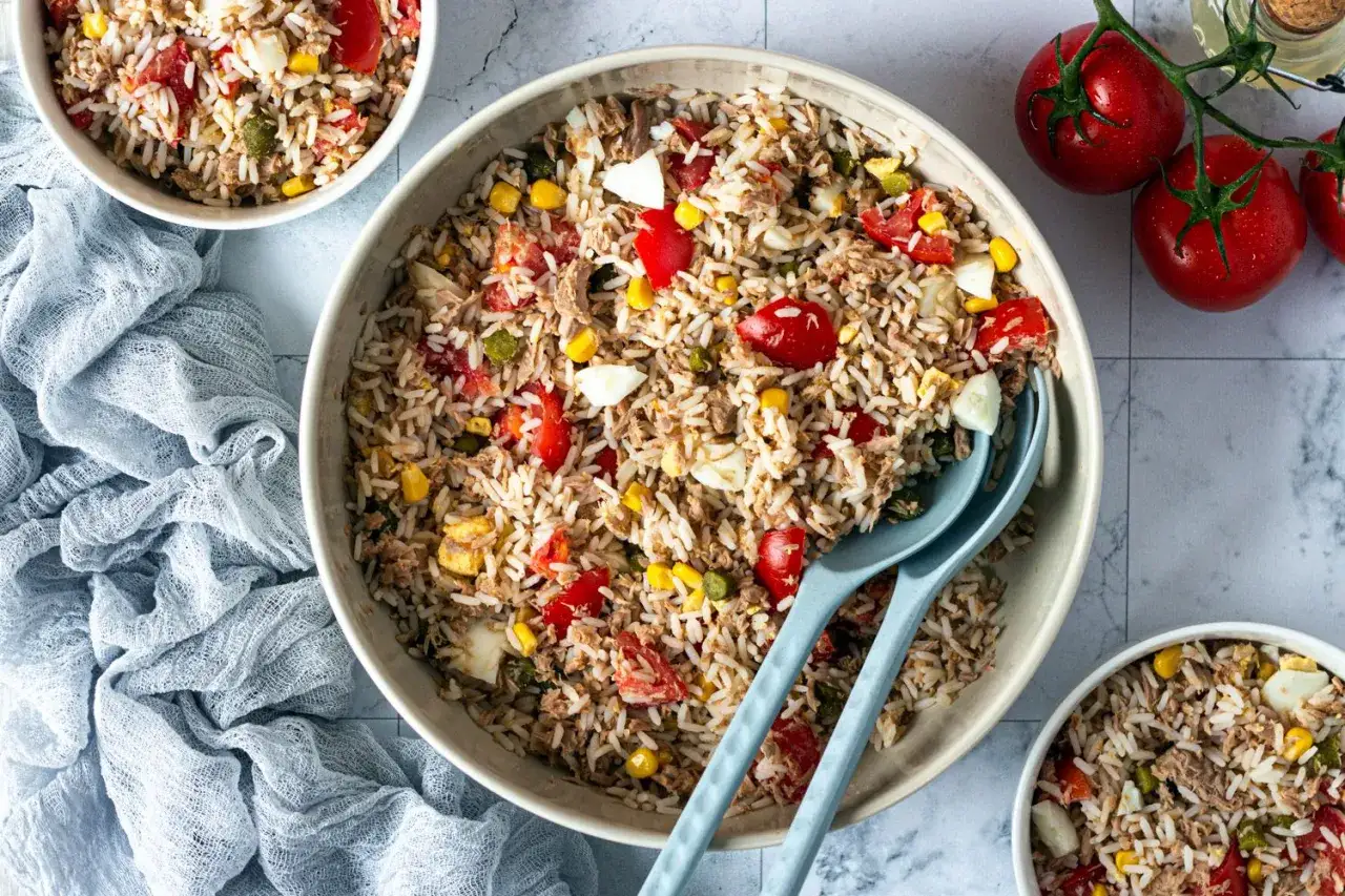 Un grand bol de salade de riz thon, garnie de tomates, maïs et œufs durs. Deux cuillères bleues sont prêtes à servir.