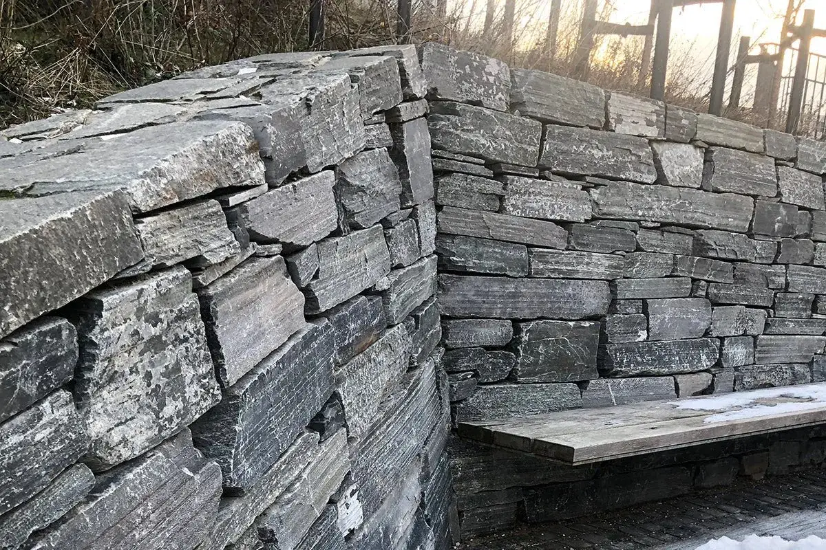 En mur av natursten som ramar in en bänk. Stenarna är grå och oregelbundna, vilket ger ett rustikt intryck.
