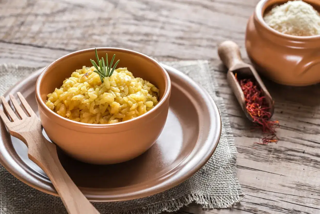 Un risotto doré, parfumé au safran, est servi dans un bol en terre cuite. À côté, une cuillère en bois révèle des pistils rouge intense.